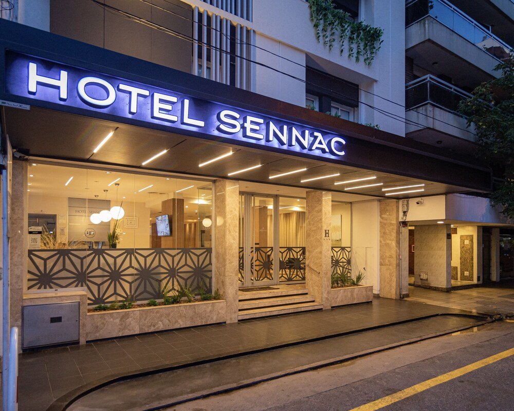 Entrada moderna del hotel con cartel iluminado, fachada de vidrio y asientos exteriores